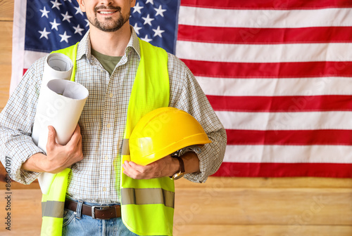 Male architect with house plans and hardhat against USA flag in workshop. Lab...