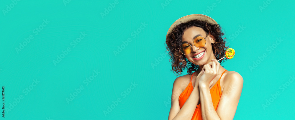 Black woman with curly brown hair smiles at the camera while holding a yellow lollipop in her right hand. She is wearing a straw hat, orange tank top, and yellow heart-shaped sunglasses. Black woman with curly brown hair smiles at the camera while holding a yellow lollipop in her right hand. She is wearing a straw hat, orange tank top, and yellow heart-shaped sunglasses.