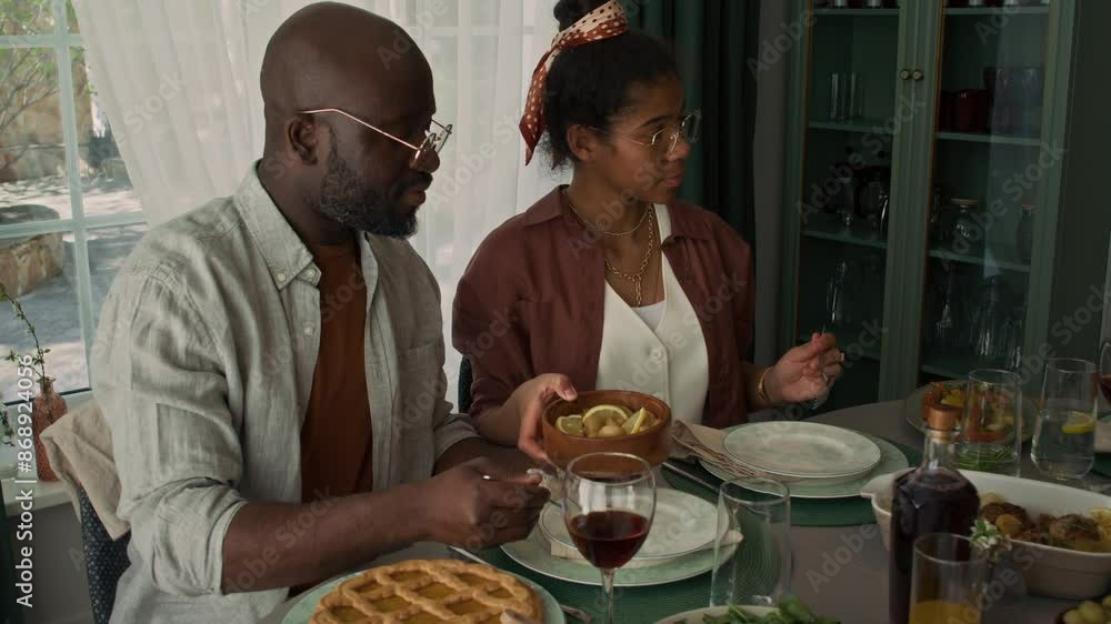 Medium shot of caring teenage African American girl passing over bowl of pickled mushrooms to father, serving spoonful on plate, while sitting at table having lunch with family Medium shot of caring teenage African American girl passing over bowl of pickled mushrooms to father, serving spoonful on plate, while sitting at table having lunch with family
