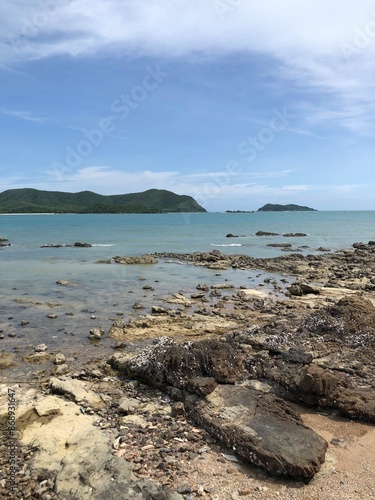 A tranquil coastal view featuring rocky shorelines and calm, turquoise waters. The sky is clear with some clouds, and green islands can be seen in the background. The foreground is dotted with barnacl