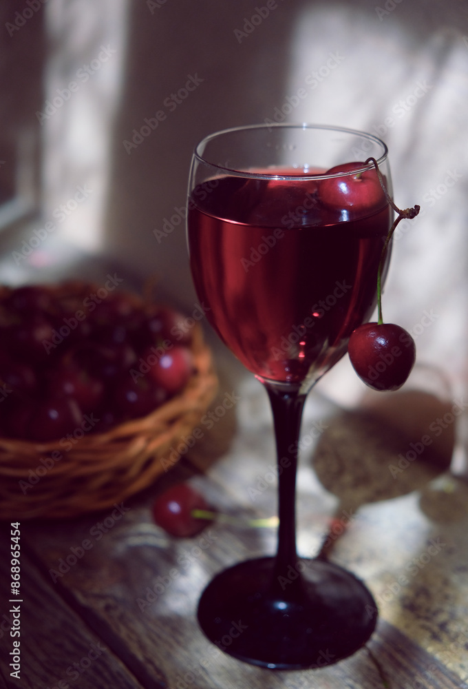 Homemade tincture of red cherry. Red sweet wine made from ripe cherries. Blurred berries in the basket. Wine glass on wooden background. Berry alcoholic drinks. In the dark, evnening atmospheric mood Homemade tincture of red cherry. Red sweet wine made from ripe cherries. Blurred berries in the basket. Wine glass on wooden background. Berry alcoholic drinks. In the dark, evnening atmospheric mood