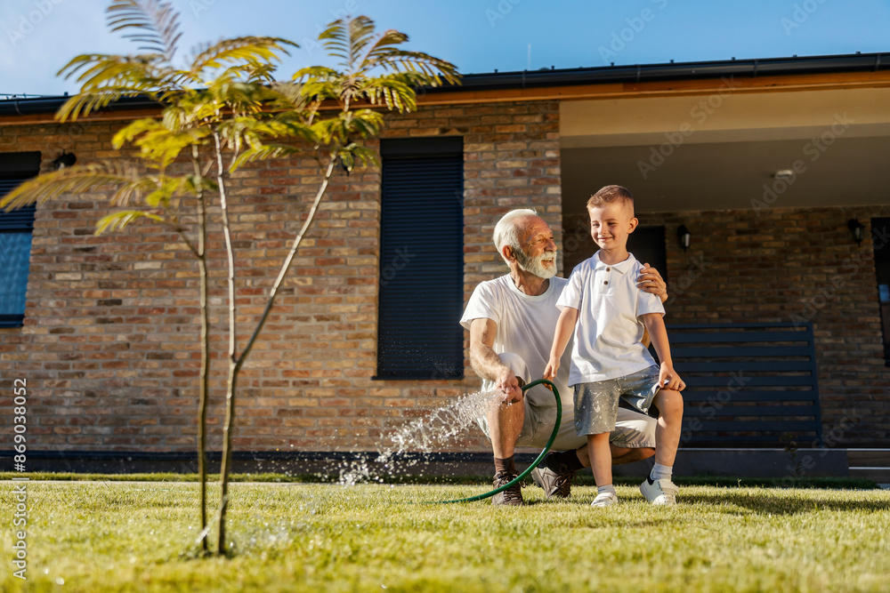 © dusanpetkovic1 - Cheerful grandson standing in backyard with his granddad hugging him while watering lawn and tree. © dusanpetkovic1 - Cheerful grandson standing in backyard with his granddad hugging him while watering lawn and tree.