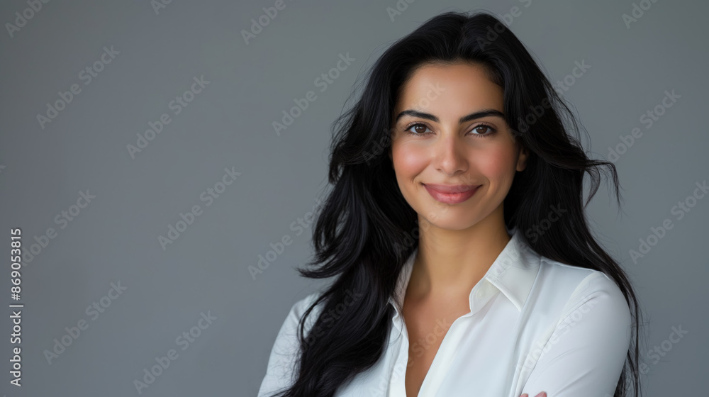 Portrait of a smiling businesswoman with Middle Eastern features, and long dark hair, standing with arms crossed against a light grey background Portrait of a smiling businesswoman with Middle Eastern features, and long dark hair, standing with arms crossed against a light grey background