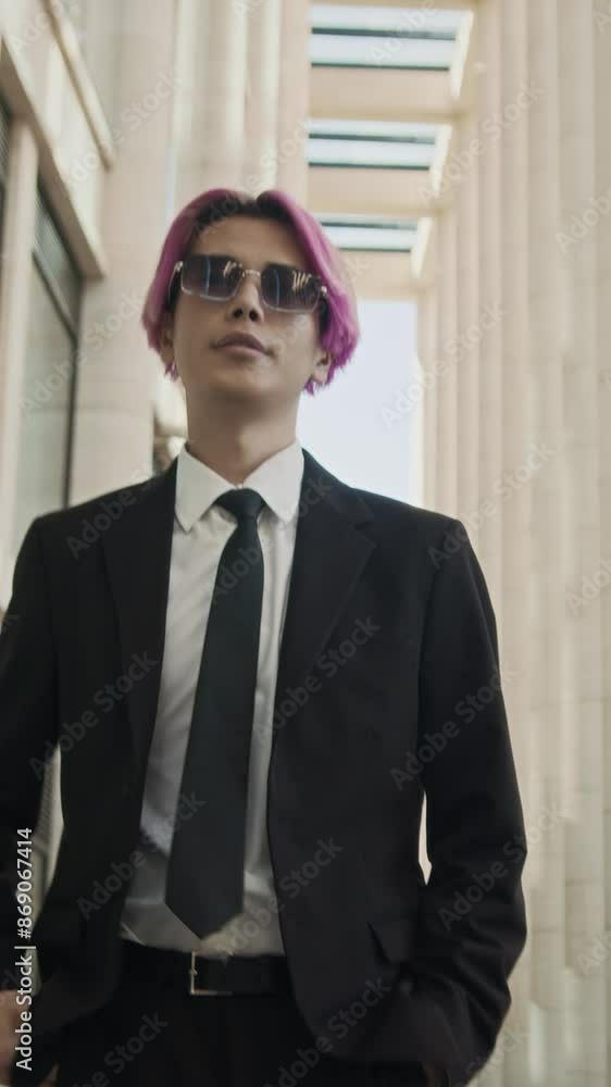 Vertical tracking medium slowmo of cool young Asian guy with pink hair wearing elegant black suit and tie and trendy glasses walking confidently down street at daytime Vertical tracking medium slowmo of cool young Asian guy with pink hair wearing elegant black suit and tie and trendy glasses walking confidently down street at daytime