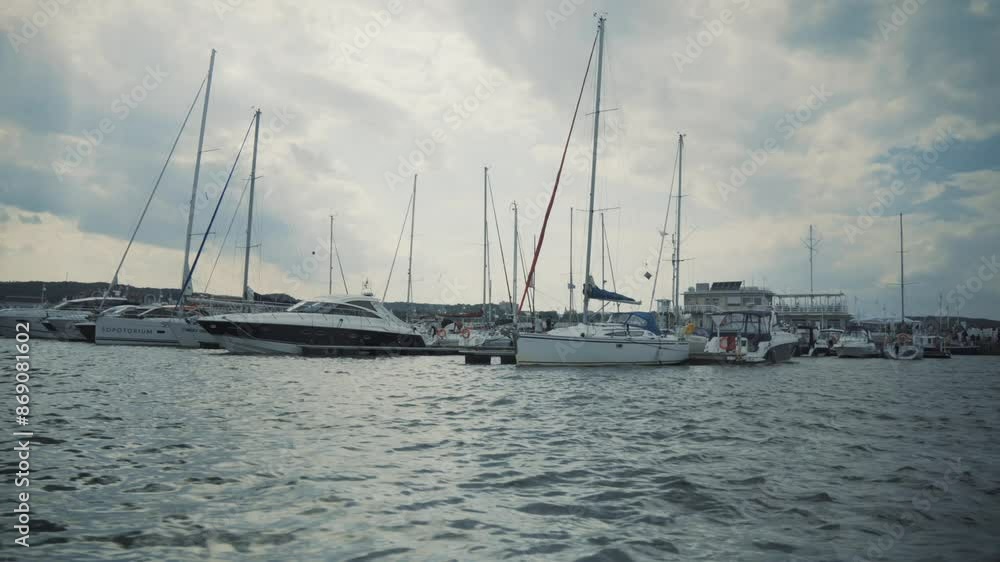 Numerous sailboats, yachts docked in a marina. Masts rising high against a partly cloudy sky. Calm water in the foreground reflects the boats. Background includes marina facilities, distant hills Numerous sailboats, yachts docked in a marina. Masts rising high against a partly cloudy sky. Calm water in the foreground reflects the boats. Background includes marina facilities, distant hills