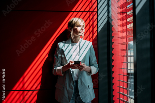 Thoughtful blond businesswoman with smart phone looking out through window at office