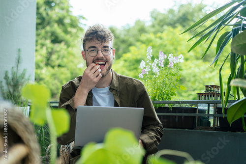 Happy young man eating cherry tomato and using laptop sitting in balcony