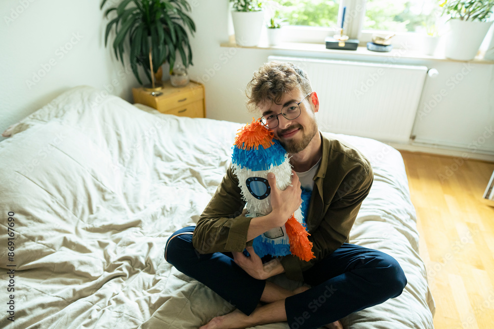 Young man hugging rocketship shaped pinata sitting on bed at home Young man hugging rocketship shaped pinata sitting on bed at home