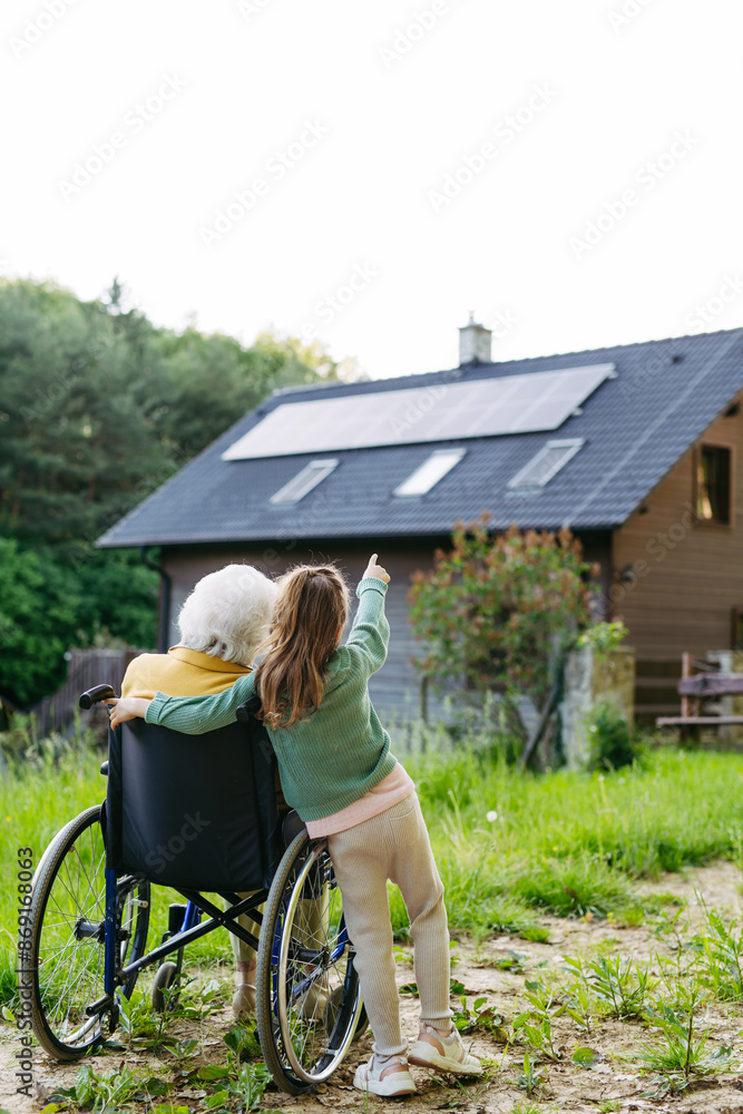 Obraz premium Granddaughter showing grandma solar panels on roof. Senior lady in wheelchair spending time in nature.