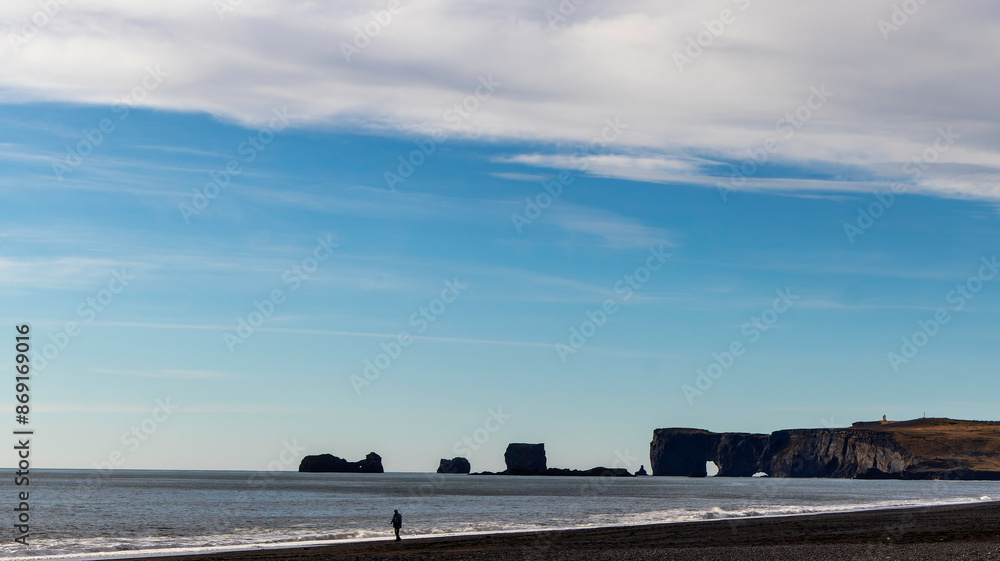 A person is walking on a beach with a view of the stone arches of Dyrholaey, Iceland A person is walking on a beach with a view of the stone arches of Dyrholaey, Iceland