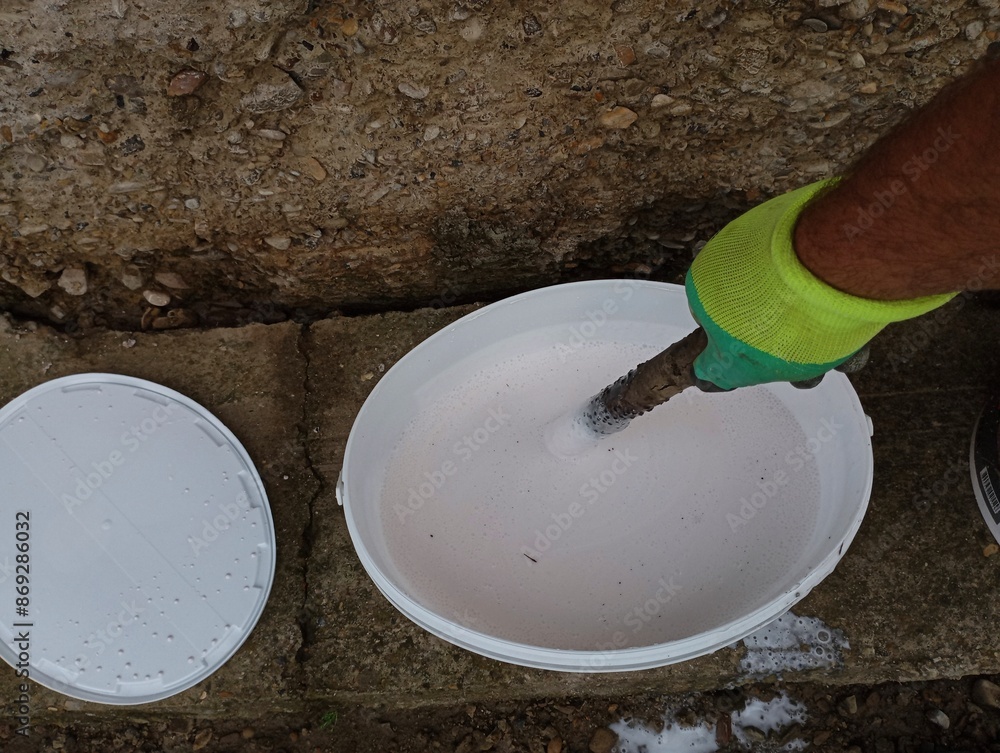 Mixing white paint in a bucket. A human hand in a work glove stirs paint in a bucket with a wooden stick. Mixing white paint in a bucket. A human hand in a work glove stirs paint in a bucket with a wooden stick.