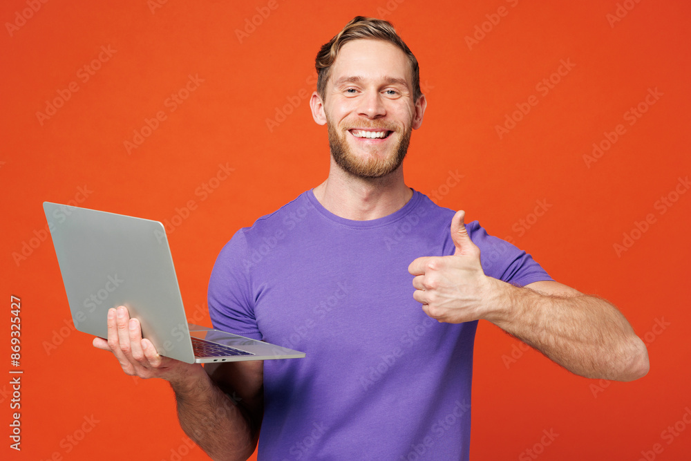 Young smiling cheerful fun happy IT man he wearing purple t-shirt casual clothes hold use work on laptop pc computer show thumb up isolated on red orange background studio portrait. Lifestyle concept. Young smiling cheerful fun happy IT man he wearing purple t-shirt casual clothes hold use work on laptop pc computer show thumb up isolated on red orange background studio portrait. Lifestyle concept.