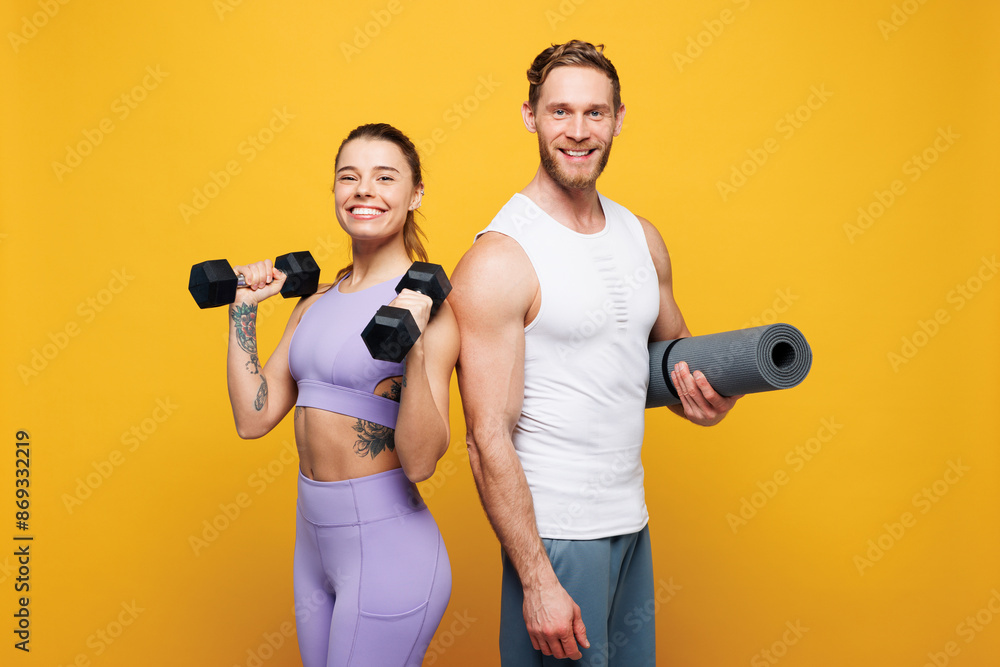 Young smiling strong fitness trainer instructor sporty two man woman wearing blue clothes spend time in home gym hold yoga mat dumbbells isolated on plain yellow background. Workout sport fit concept. Young smiling strong fitness trainer instructor sporty two man woman wearing blue clothes spend time in home gym hold yoga mat dumbbells isolated on plain yellow background. Workout sport fit concept.