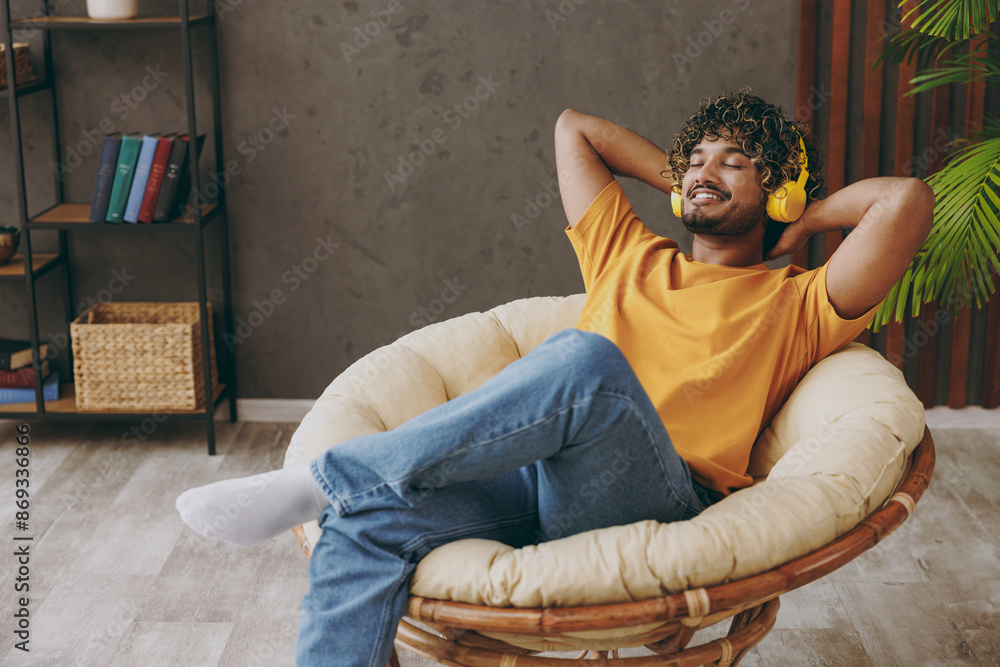 © ViDi Studio - Young happy cool Indian man he wear orange casual clothes listen to music in headphones sits in armchair stay at home hotel flat rest relax spend free spare time in living room indoor. Lounge concept.