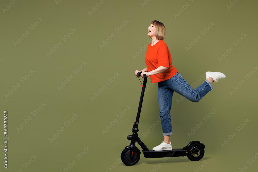 Full body side profile view elderly blonde woman 50s years old wears orange t-shirt casual clothes riding electric scooter isolated on plain pastel green background studio portrait. Lifestyle concept. Full body side profile view elderly blonde woman 50s years old wears orange t-shirt casual clothes riding electric scooter isolated on plain pastel green background studio portrait. Lifestyle concept.