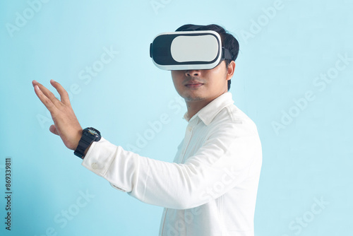 Isolated man wearing VR headset and white shirt, standing and gesture with hand, fully immersed in a virtual reality experience, interacting with a virtual environment on light blue background