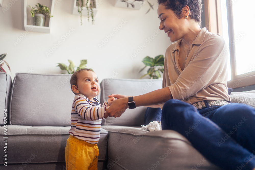 Joyful baby boy holding hands with his smiling mother while learning to walk. Adorable toddler in colorful striped outfit receiving support and encouragement at home.. Joyful baby boy holding hands with his smiling mother while learning to walk. Adorable toddler in colorful striped outfit receiving support and encouragement at home..