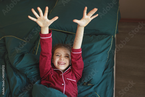 Happy 7 year old girl in red pyjamas in green blanket arms outstretched on bed at home photo