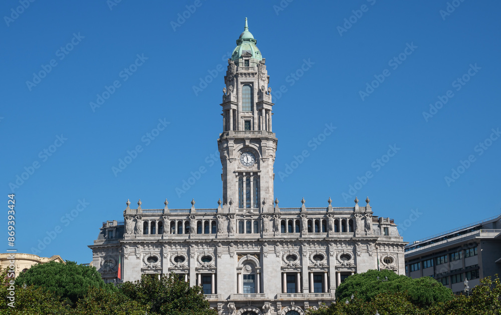 Naklejka premium Elegant historical building with a prominent clock tower, ornate sculptures, and baroque architectural details stands under a clear blue sky, flanked by lush greenery in a serene urban setting.