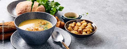 Bowl of mashed potato soup with a spoon on the plate. The bowl is filled with yellow soup puree and topped with croutons, parsley and ground red pepper.