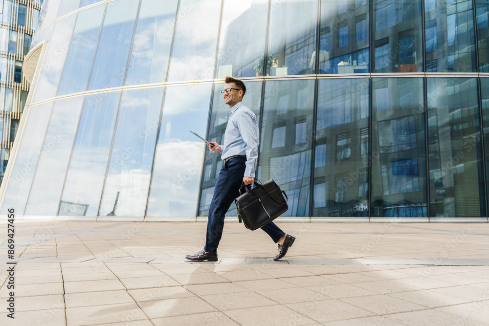 Smiling businessman in blue shirt walking with a briefcase and tablet, near modern glass buildings. Smiling businessman in blue shirt walking with a briefcase and tablet, near modern glass buildings.