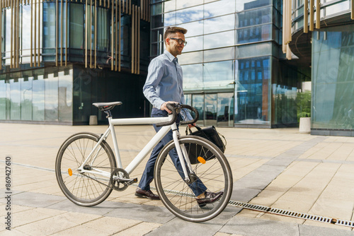 Smiling professional man with glasses, walking a white bicycle and holding a briefcase near glass office building.