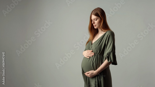 A pregnant woman in a green dress poses, holding her hands on her belly, standing against a neutral background.
