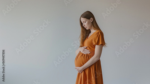 A pregnant woman in an orange dress poses, holding her hands on her belly, standing against a neutral background.
