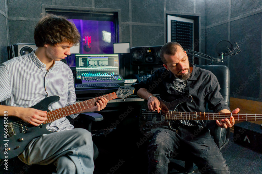 Obraz premium in the equipment of the recording room handsome man with a beard and tattoos and his young friend play the guitar in pairs and act out a song
