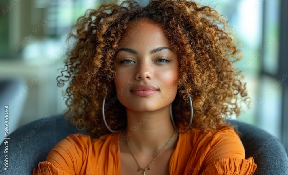 Fototapeta premium A woman with curly hair and a gold necklace is sitting in a chair. She has a smile on her face and is looking directly at the camera