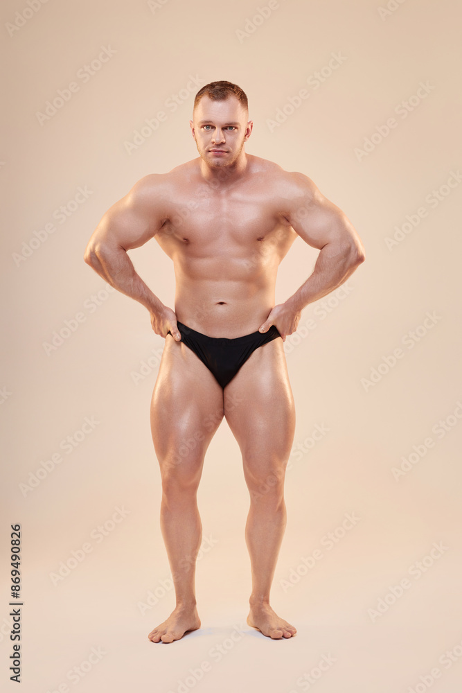 Full shot portrait of topless muscular bodybuilder in front lat spread pose with hands on waist Full shot portrait of topless muscular bodybuilder in front lat spread pose with hands on waist