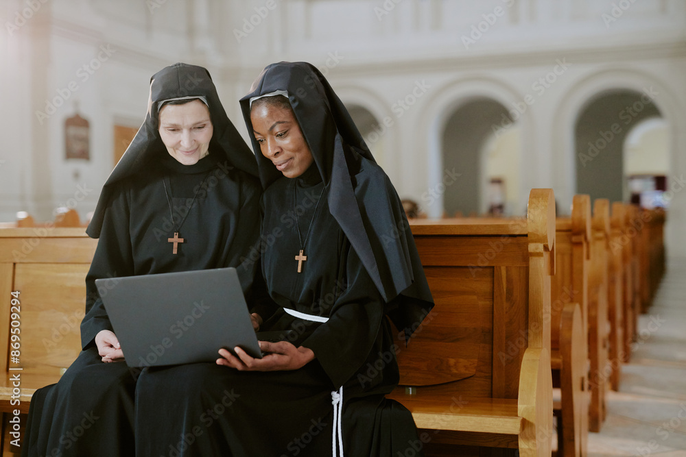 Medium shot of two ethnically diverse young and mature nuns sitting on wooden bench in Catholic church working on laptop Medium shot of two ethnically diverse young and mature nuns sitting on wooden bench in Catholic church working on laptop