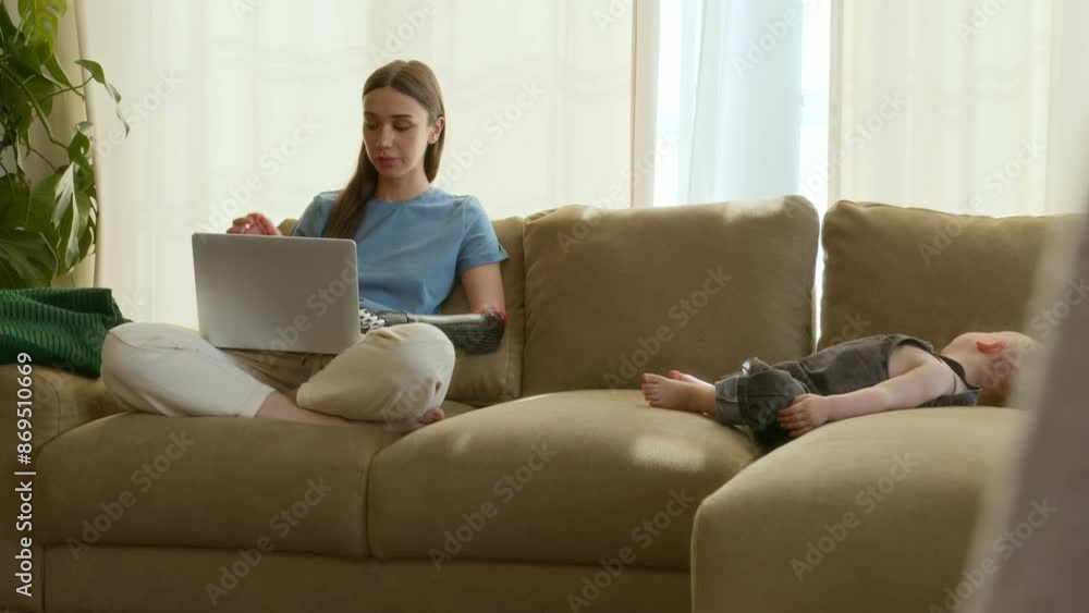 Young mother with prosthetic arm is working remotely on laptop while baby is napping on couch in living room. Female freelance worker uses bionic arm to type on keyboard Young mother with prosthetic arm is working remotely on laptop while baby is napping on couch in living room. Female freelance worker uses bionic arm to type on keyboard