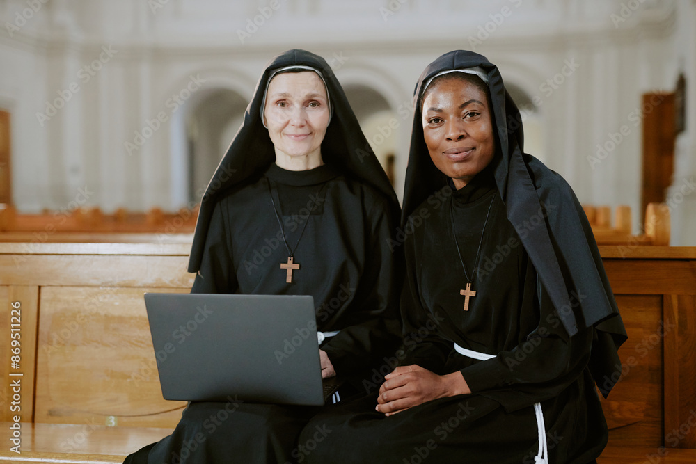 Medium shot of two ethnically diverse nuns sitting on wooden pew in Catholic church, looking at camera while using laptop Medium shot of two ethnically diverse nuns sitting on wooden pew in Catholic church, looking at camera while using laptop