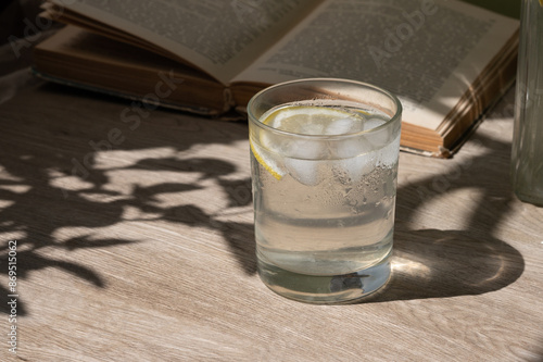 Glass with iced refreshing lemonade drink or lemon infused water, open book on wooden table background with aesthetic floral sunlight shadows, healthy summer beverage, copy space photo