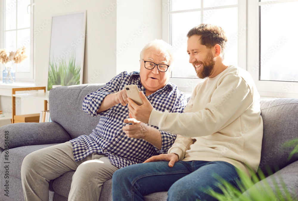 Old father and adult son sitting on sofa looking photos on phone laughter and smiling at whole room. Old father and adult son family moments happiness and love father and son looking photos on phone Old father and adult son sitting on sofa looking photos on phone laughter and smiling at whole room. Old father and adult son family moments happiness and love father and son looking photos on phone