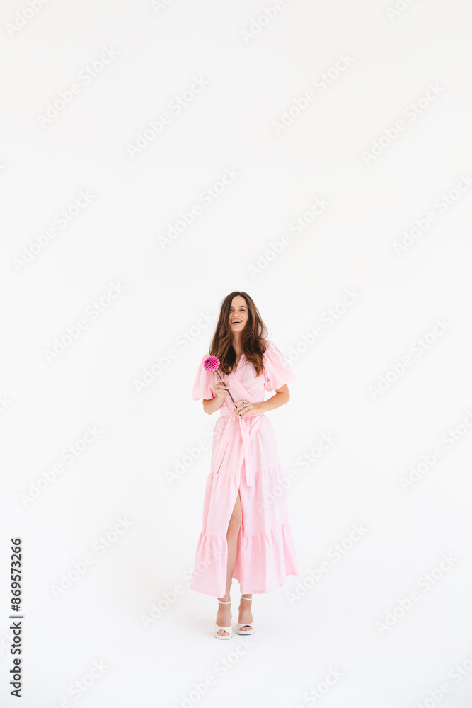 Model wearing casual outfit. Look in trendy minimalistic style. Pink dress with slit, belt, puff sleeves. Sexy sensual happy woman. Positive woman turn around and hold pink flower on white background. Model wearing casual outfit. Look in trendy minimalistic style. Pink dress with slit, belt, puff sleeves. Sexy sensual happy woman. Positive woman turn around and hold pink flower on white background.