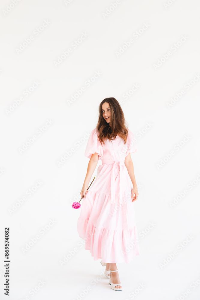 Model wearing casual outfit. Look in trendy minimalistic style. Pink dress with slit, belt, puff sleeves. Sexy sensual happy woman. Positive woman turn around and hold pink flower on white background. Model wearing casual outfit. Look in trendy minimalistic style. Pink dress with slit, belt, puff sleeves. Sexy sensual happy woman. Positive woman turn around and hold pink flower on white background.