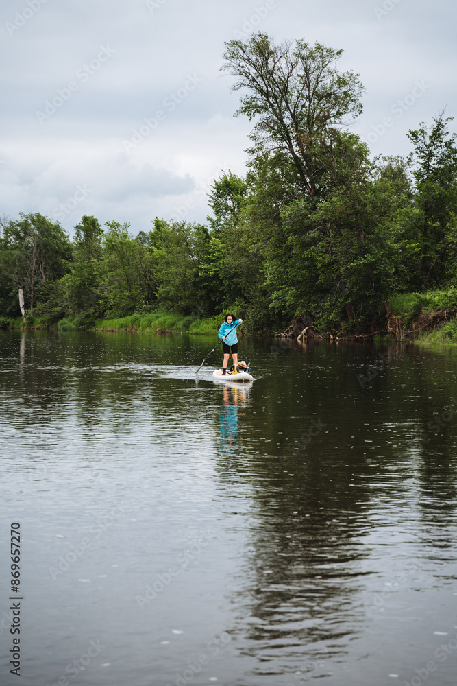 In a serene scene, a man peacefully paddles on a board in a calm river, captivated by the beauty of the water, sky, clouds, and trees around him, creating a tranquil moment of serenity In a serene scene, a man peacefully paddles on a board in a calm river, captivated by the beauty of the water, sky, clouds, and trees around him, creating a tranquil moment of serenity