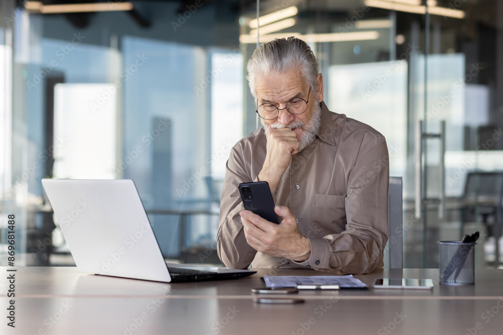Senior businessman in modern office, deeply engaged with his smartphone while working on a laptop. The image conveys concentration, modern technology, and professional work environment. Senior businessman in modern office, deeply engaged with his smartphone while working on a laptop. The image conveys concentration, modern technology, and professional work environment.