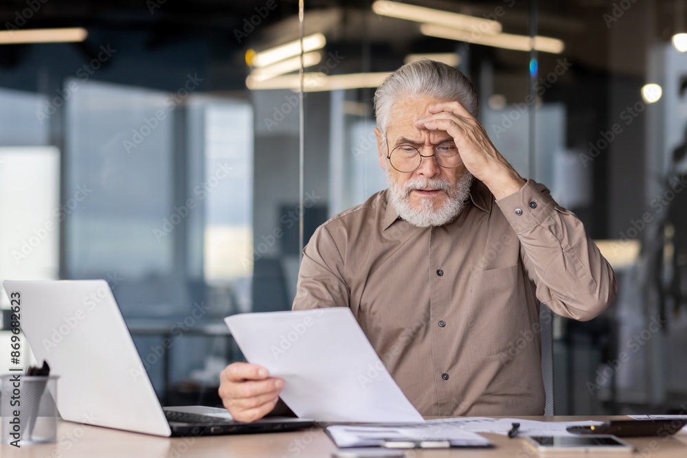 Senior businessman looking stressed while reading documents at his office desk. He is sitting in front of a laptop, holding a paper and appears to be in deep thought or confusion. Senior businessman looking stressed while reading documents at his office desk. He is sitting in front of a laptop, holding a paper and appears to be in deep thought or confusion.