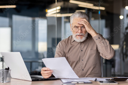 Wallpaper Mural Senior businessman looking stressed while reading documents at his office desk. He is sitting in front of a laptop, holding a paper and appears to be in deep thought or confusion. Torontodigital.ca
