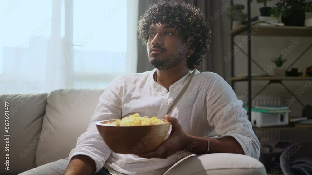 Portrait of tired young curly man with bowl of popcorn sitting on sofa and watching boring uninteresting movie show series while looking at screen at cozy home alone Portrait of tired young curly man with bowl of popcorn sitting on sofa and watching boring uninteresting movie show series while looking at screen at cozy home alone
