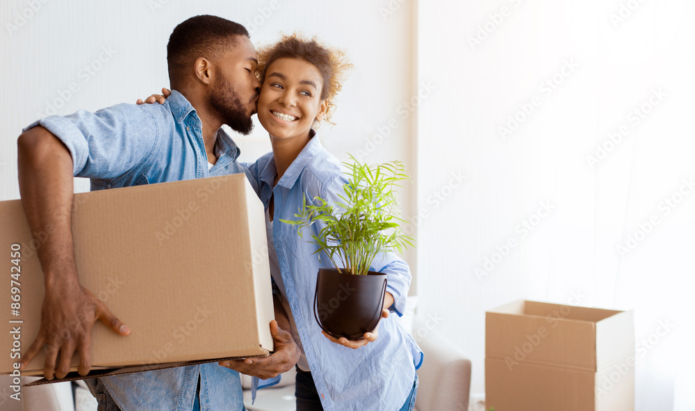 Afro Husband Kissing Wife In Cheek Holding Moving Box Unpacking In New Apartment. Empty Space Afro Husband Kissing Wife In Cheek Holding Moving Box Unpacking In New Apartment. Empty Space