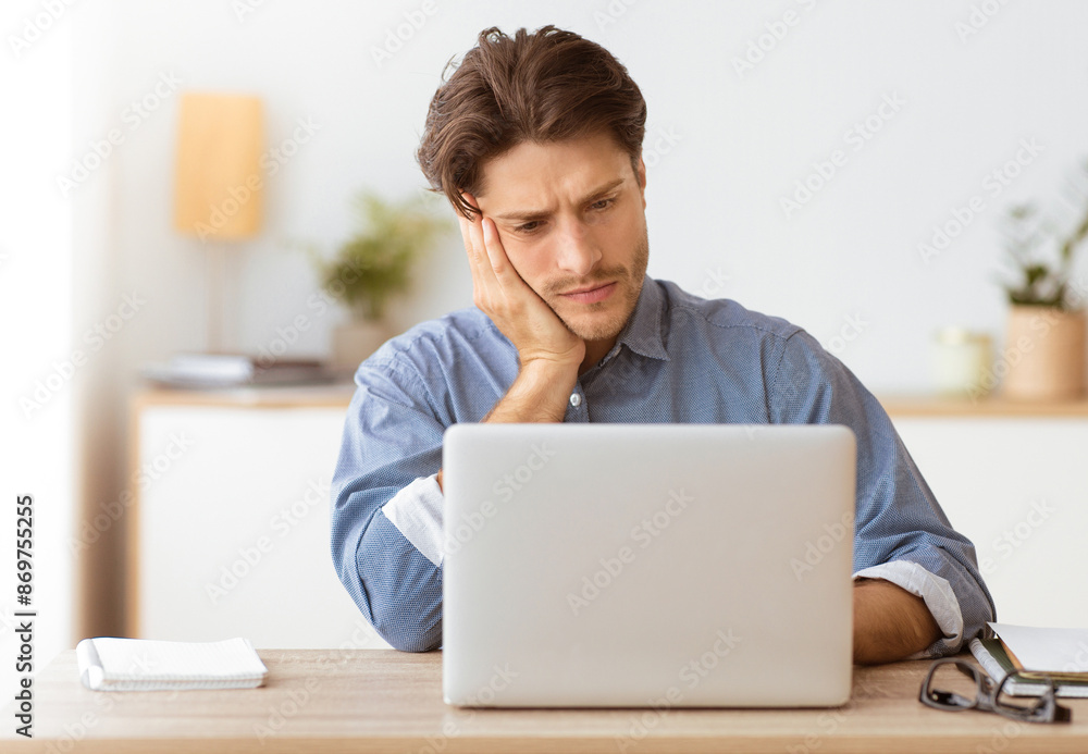A man sits at a desk with a laptop in front of him, looking concerned. He rests his head on his hand while looking at the screen. A man sits at a desk with a laptop in front of him, looking concerned. He rests his head on his hand while looking at the screen.