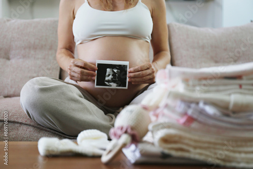 Pregnant woman with big belly preparing for the arriving of a newborn, folding child's clothing. Expecting female on a third trimester withultrasound image of child. Close up, copy space, background.