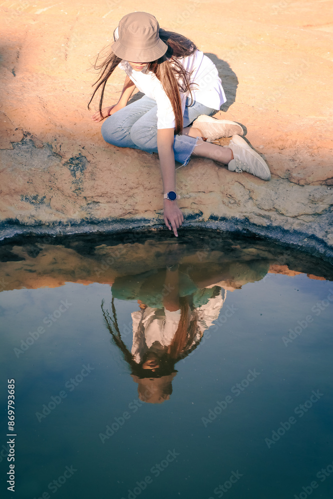 A teenage girl in a Panama hat with flowing long hair sits cross-legged on a rocky shore, runs her hand through the water and looks at her reflection in the water. A teenage girl in a Panama hat with flowing long hair sits cross-legged on a rocky shore, runs her hand through the water and looks at her reflection in the water.