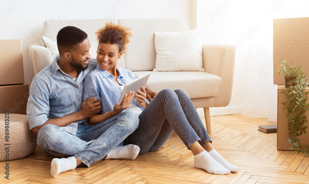 Relocation Concept. Joyful Black Spouses Using Tablet Computer Sitting On Floor Among Moving Boxes In New House. Copy Space Relocation Concept. Joyful Black Spouses Using Tablet Computer Sitting On Floor Among Moving Boxes In New House. Copy Space