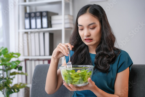 фотографія Young woman making disgusted face while eating salad at home