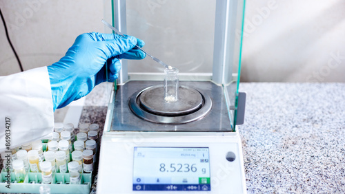 A scientist's gloved hand uses a stainless spatula to transfer a substance into a vial for weighing a sample with a four-decimal digital analytical balance with 4-digit precision in the laboratory.